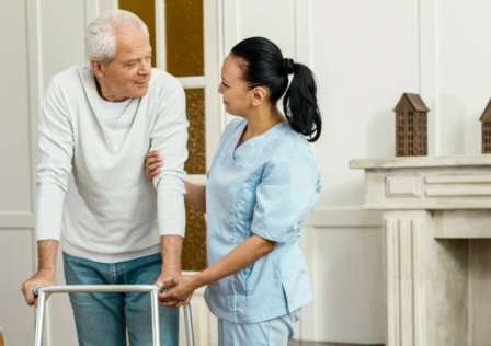 professional caregiver standing near her patient and holding his hand while helping him to walk